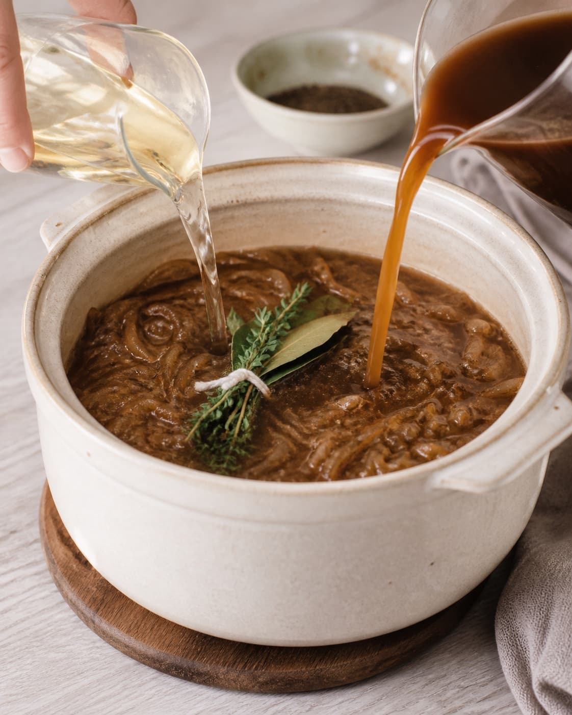 Caramelized onions becoming clear brown soup as white wine and beef stock are added, tied thyme-bay bundle visible, no bread or cheese under tilberedningen af løgsuppe