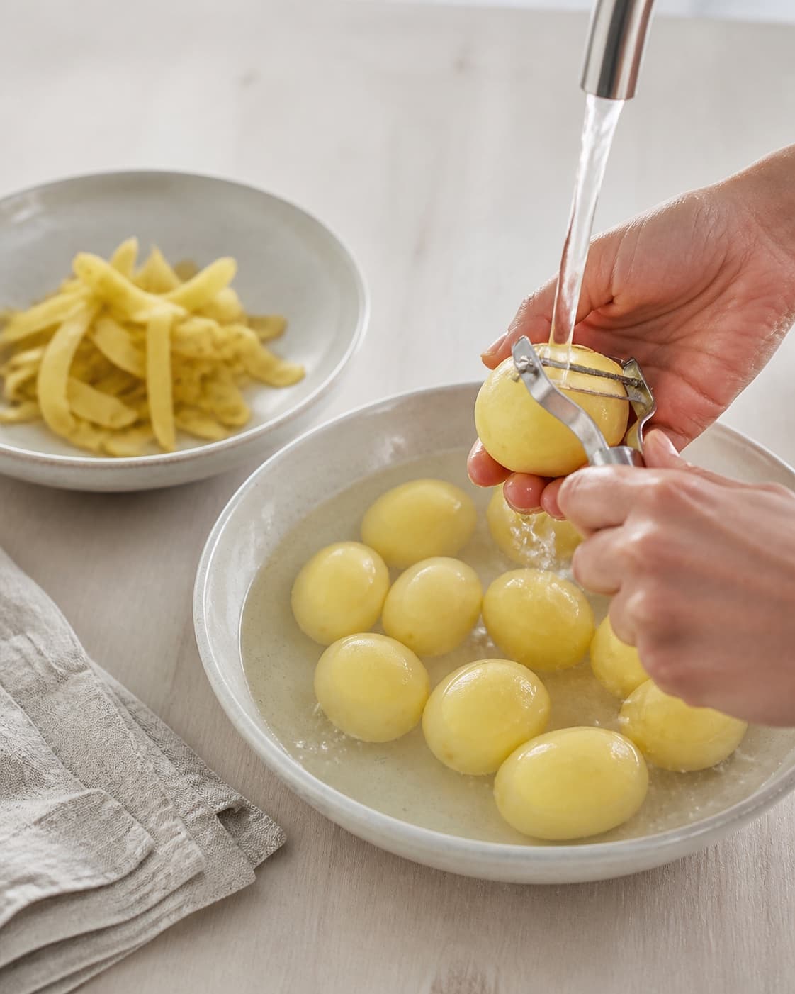 Fresh potatoes being thinly peeled and rinsed, smooth pale-yellow surfaces with no eyes or dark spots, no cooking pot under tilberedningen af kogte kartofler