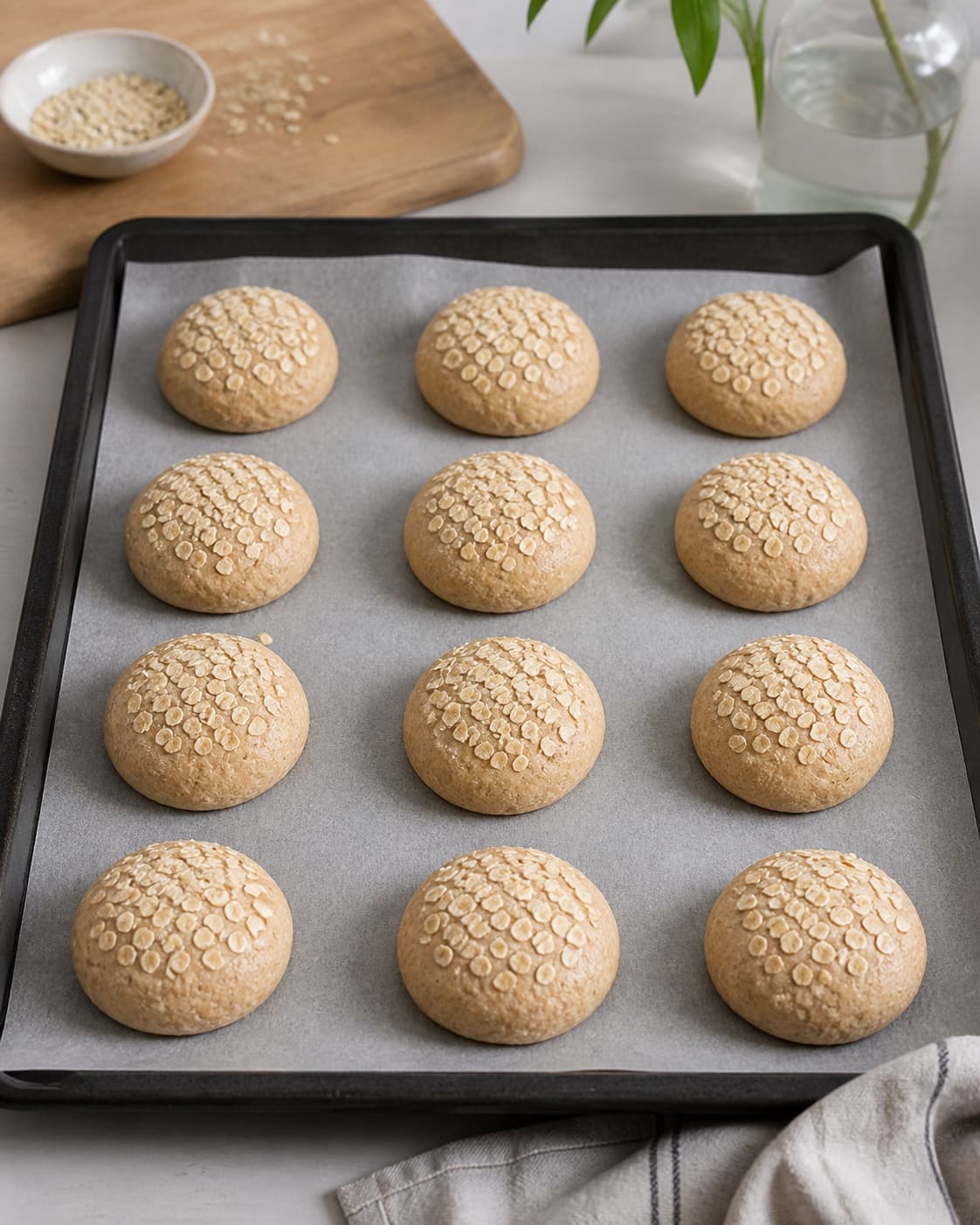 Exactly twelve equal raw rye rolls arranged with gaps on one baking tray, each topped with rolled oats, no baked rolls under tilberedningen af rugboller