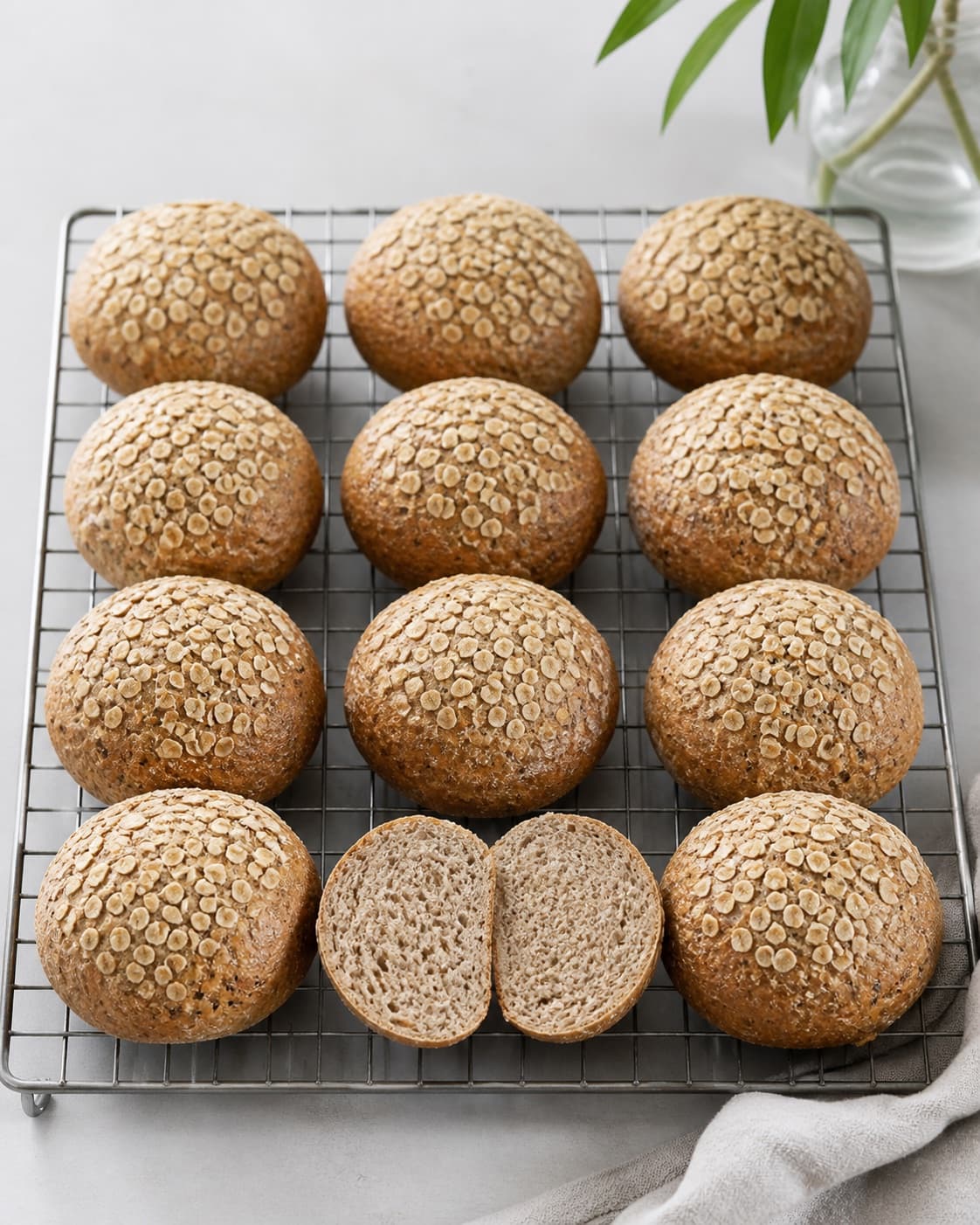 Exactly twelve golden-brown rye rolls with oat topping cooling on one rack, one split open showing moist coarse crumb, no extra rolls under tilberedningen af rugboller