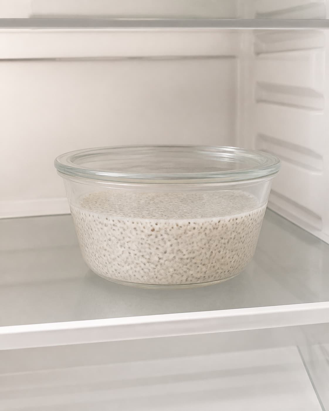 One covered clear glass bowl of fully set cold coconut chia pudding alone on a clean refrigerator shelf, no fruit nuts yoghurt or extra containers under tilberedningen af kokos-chiagrød