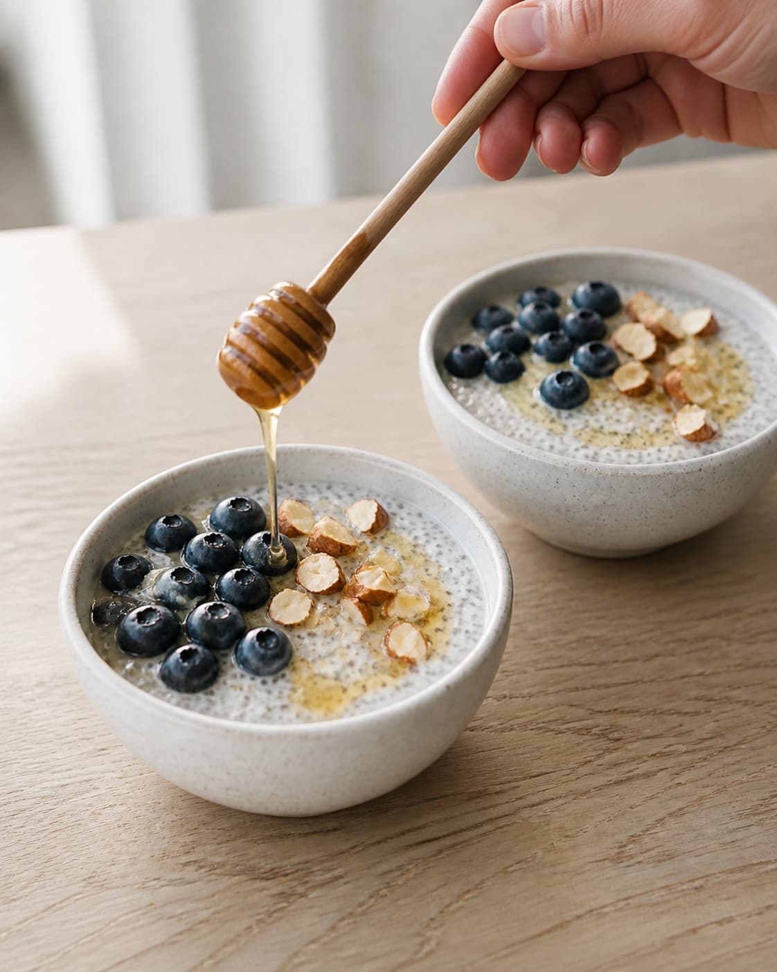 Exactly two bowls of creamy coconut chia pudding topped with blueberries, coarsely chopped almonds and a thin honey drizzle, no extra bowls cups books or eggs under tilberedningen af kokos-chiagrød