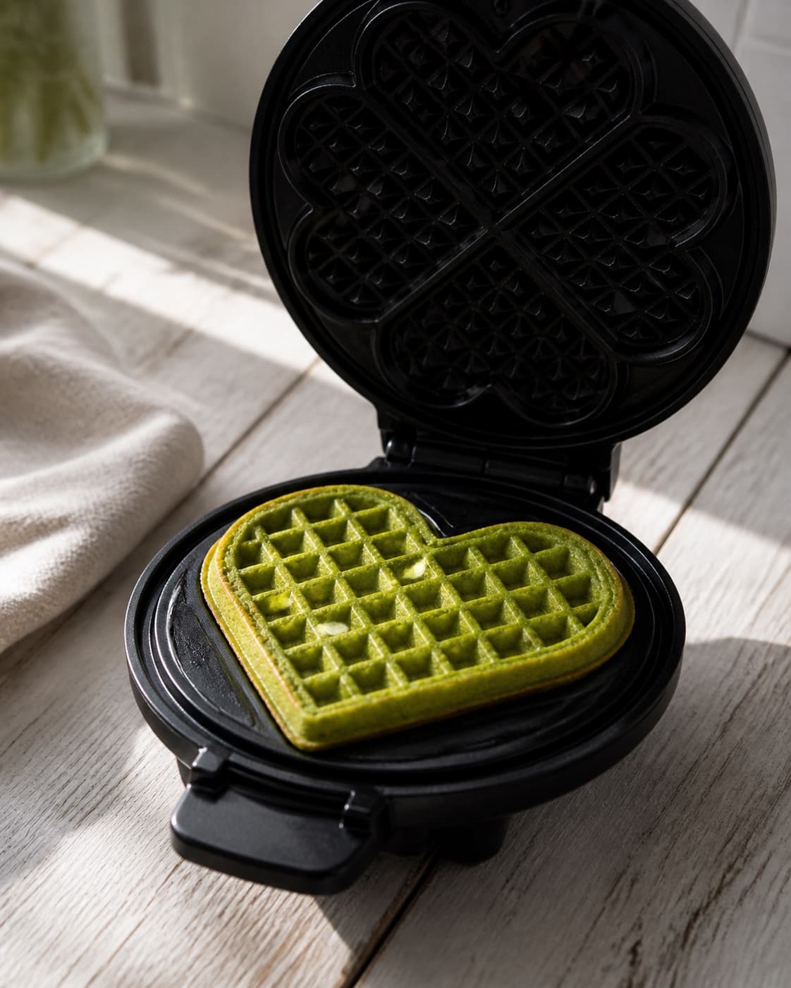 One open heart-shaped waffle iron holding one vivid-green cooked waffle with crisp golden edges, no roe cream onion dill or extra waffles under tilberedningen af grønkålsvafler med stenbiderrogn