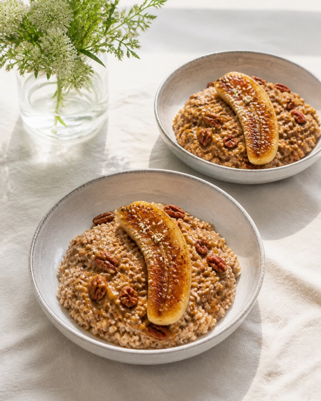 Exactly two bowls of spiced buckwheat porridge, each with one caramelized banana half, chopped pecans, hemp seeds and a small almond-butter drizzle, no extra bowls cups or whole bananas under tilberedningen af boghvedegrød med karamelliseret banan