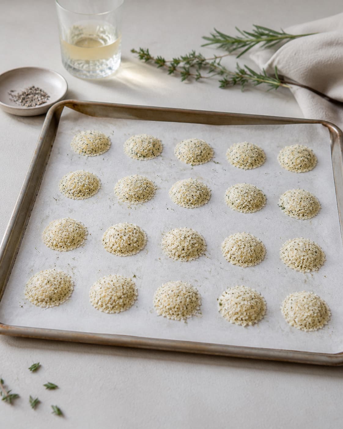Exactly twenty small round mounds of grated herb Parmesan in four rows of five on one parchment-lined tray, no melted or baked chips under tilberedningen af parmesanchips med krydderurter