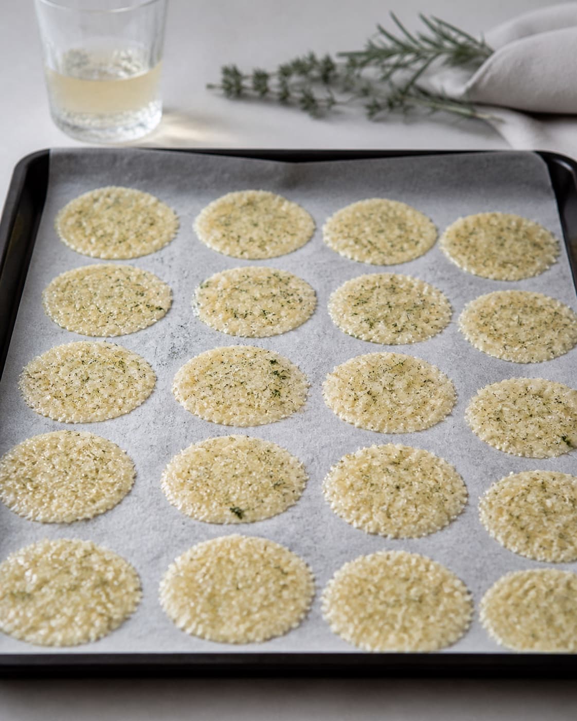Twenty pale-golden round Parmesan crisps melting on parchment in one oven tray, no dark brown edges or extra food under tilberedningen af parmesanchips med krydderurter