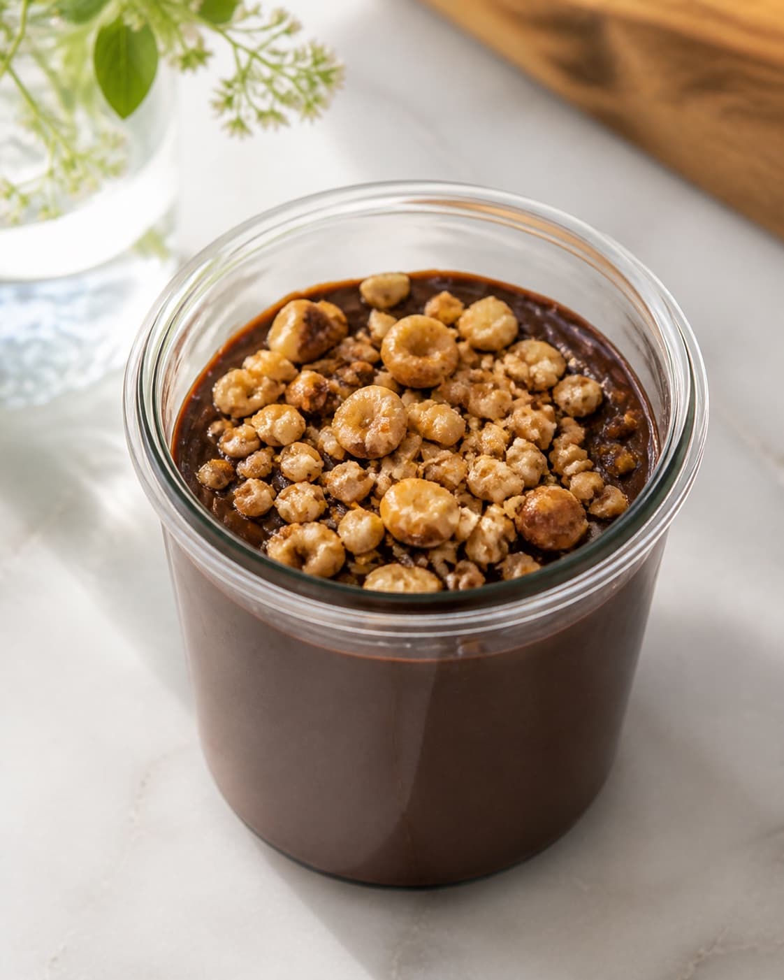 One single open glass jar filled with glossy dark chocolate spread and topped with a clear layer of golden hazelnut crunch, no bread extra jars or loose ingredients under tilberedningen af chokoladesmør med hasselnøddeknas