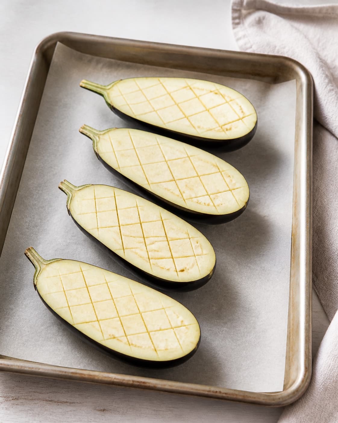 Exactly four raw aubergine halves on one baking tray, pale flesh deeply scored in a clear diamond grid, no marinade bulgur or toppings under tilberedningen af bagte auberginer med bulgur