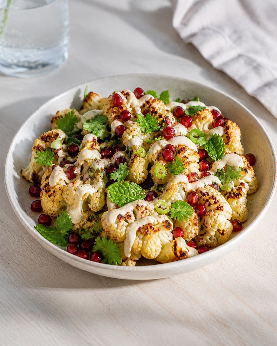 One finished platter of golden cauliflower drizzled with pale tahini dressing and topped with red pomegranate seeds and fresh green herbs, no extra plates under tilberedningen af stegt blomkål med tahin og granatæble