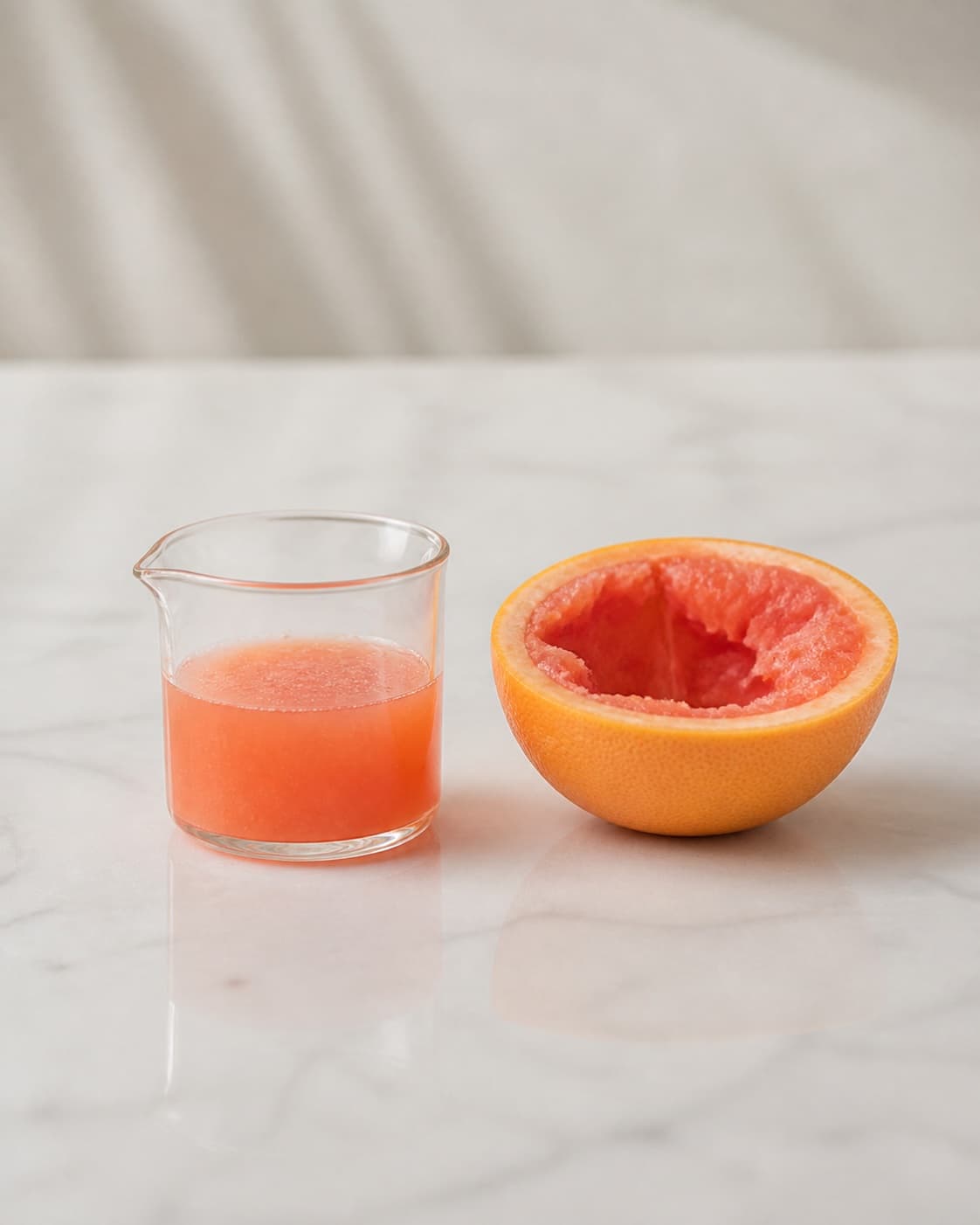 One small measuring glass holding pink freshly squeezed grapefruit juice beside one squeezed grapefruit half, no gin mint pineapple ice or cocktail glass under tilberedningen af summer breeze cocktail