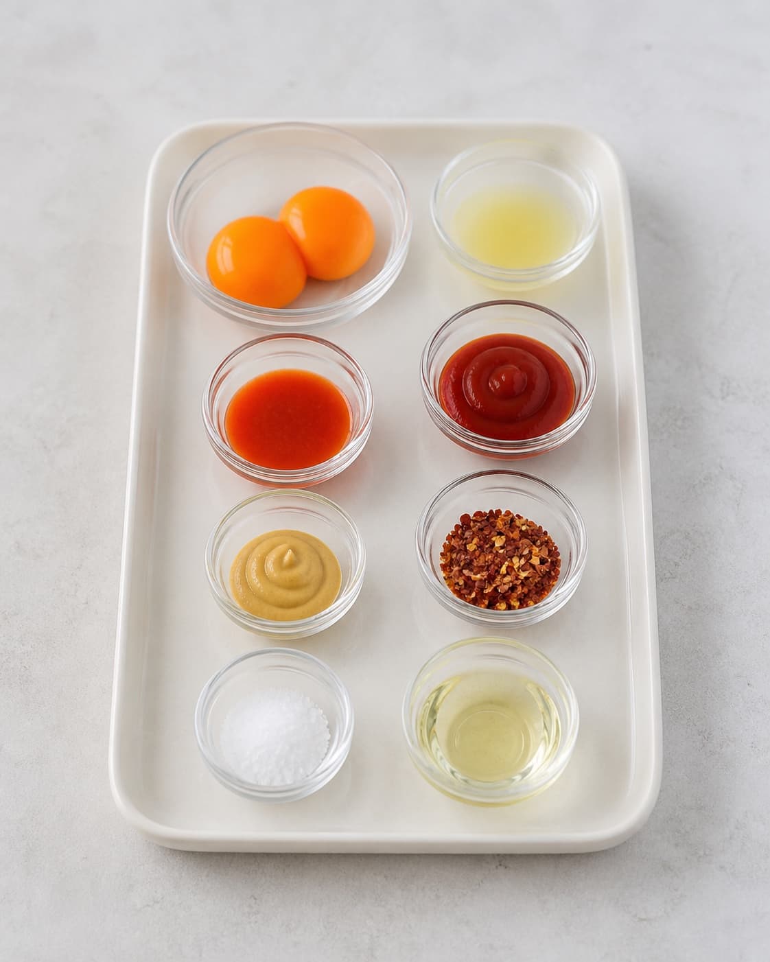 One clean tray holding two egg yolks and small bowls of lemon juice sriracha ketchup Dijon and oil, no finished mayonnaise blender or garnish under tilberedningen af chilimayo