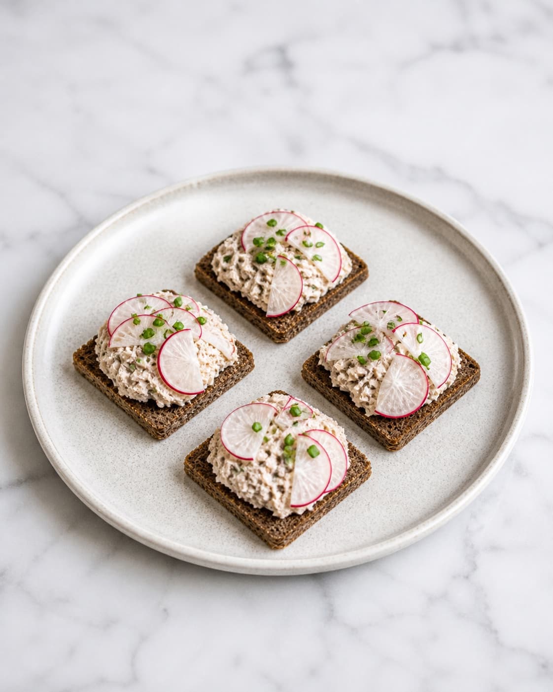 Exactly four small pieces of toasted rye bread topped with smoked mackerel mousse, radish slices and chives on one platter, no extra sandwiches bowls or garnish under tilberedningen af makrelmousse - røget makrelrilette
