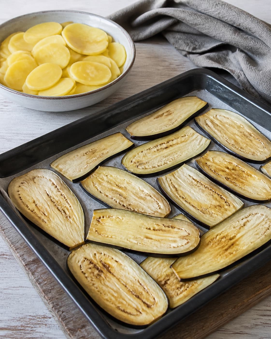 One baking tray of pliable lightly golden aubergine slices beside one bowl of thin parboiled potato slices, no sauce cheese meat or garnish under tilberedningen af moussaka med kartofler og aubergine