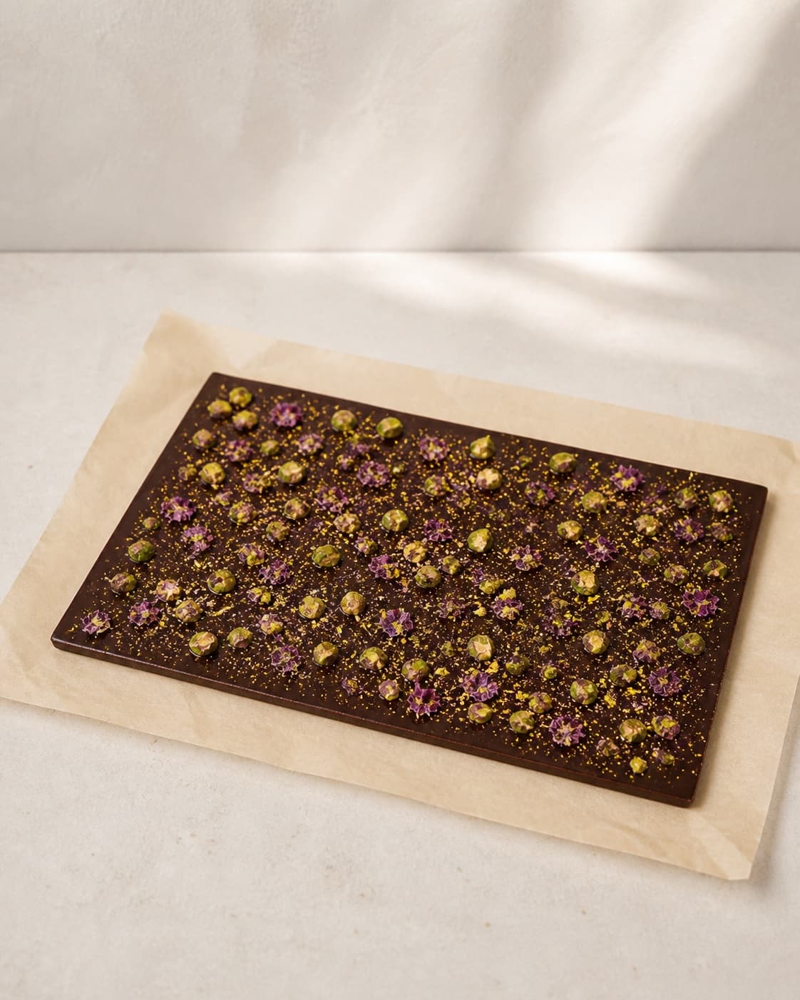One unset flat glossy dark chocolate slab on parchment evenly topped with chopped pistachios, sparse edible dried flowers and a tiny dusting of gold, no broken pieces under tilberedningen af chokoladebrud med pistaciekerner