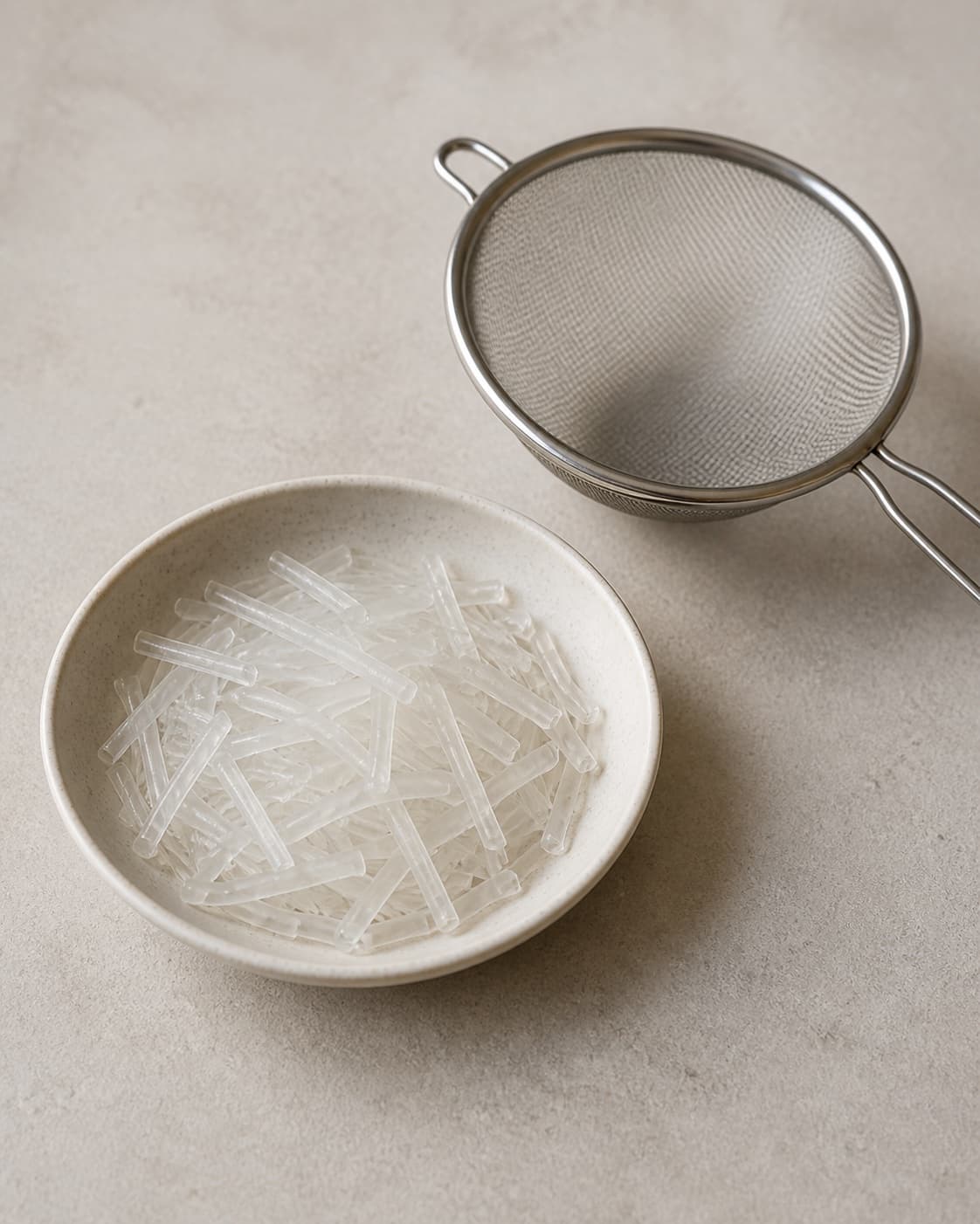 One bowl holding clear softened short glass noodles beside one strainer, no meat vegetables wrappers sauce or garnish under tilberedningen af sprøde forårsruller