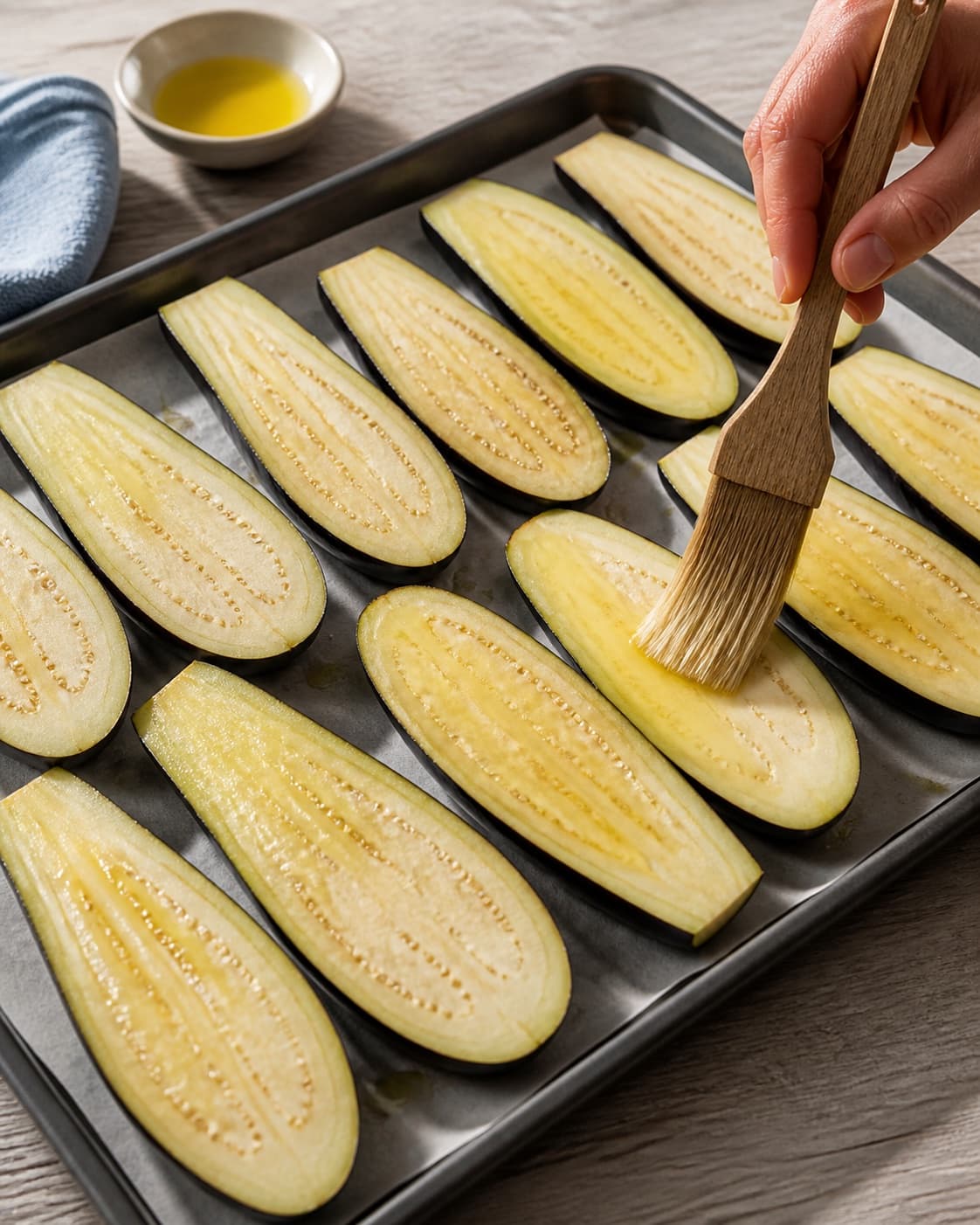 One baking tray holding evenly cut raw lengthwise aubergine slices being brushed glossy with olive oil, no dressing nuts herbs pomegranate or garnish under tilberedningen af grillet aubergine med tahindressing og mynte