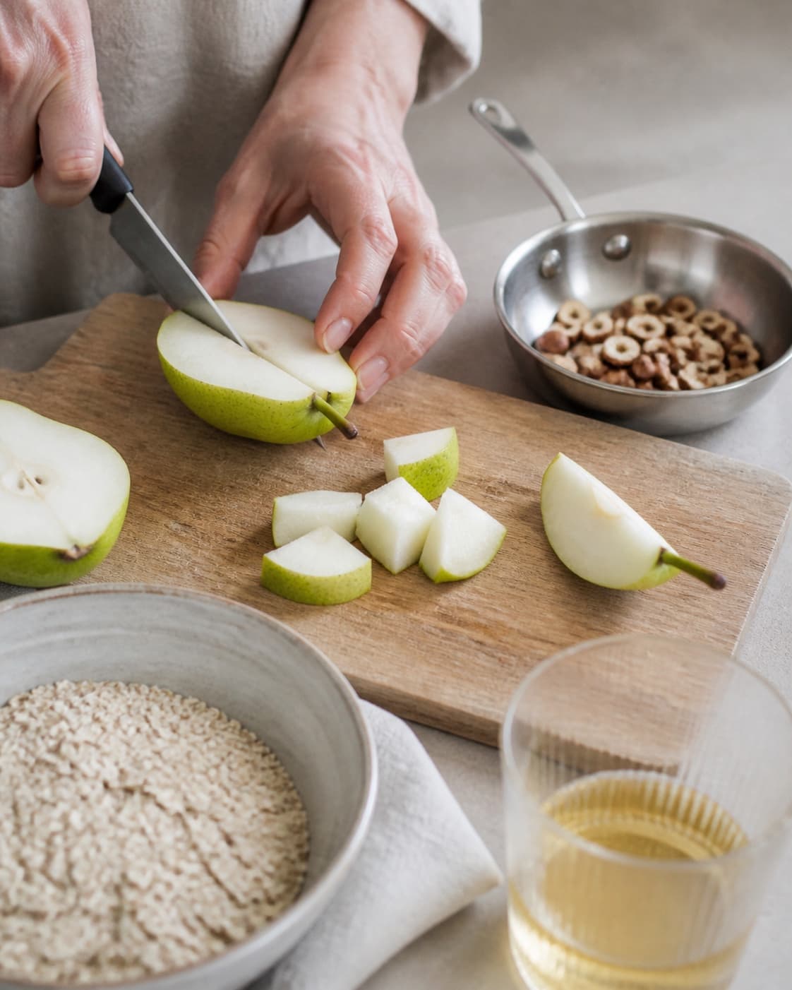 One single photorealistic instructional frame of fresh fruit being cut beside one small pan of toasted topping for Bircher Müsli med pære, featuring grove havregryn, pære, æblejuice, with only the named equipment and no unrelated food, text, logos or packaging under tilberedningen af bircher müsli med pære