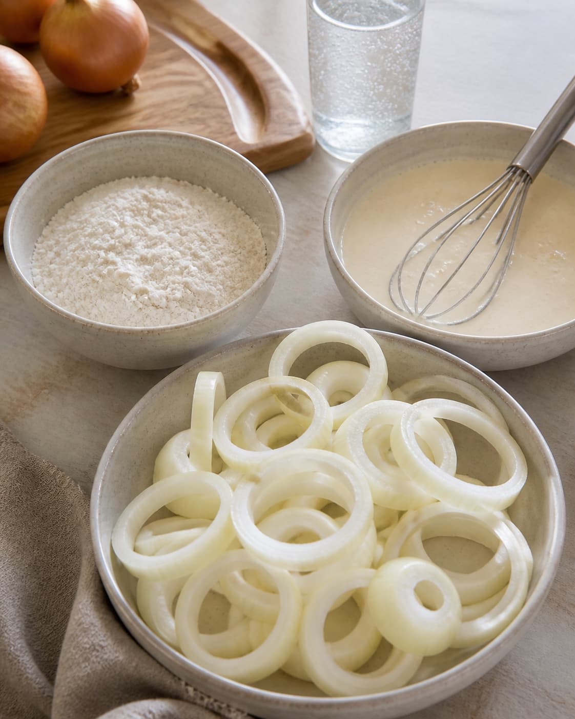 One single photorealistic instructional frame of uniform raw pieces prepared for breading for Sprøde løgringe med chilicheese dip, featuring store løg, hvedemel, danskvand, with only the named equipment and no unrelated food, text, logos or packaging under tilberedningen af sprøde løgringe med chilicheese dip