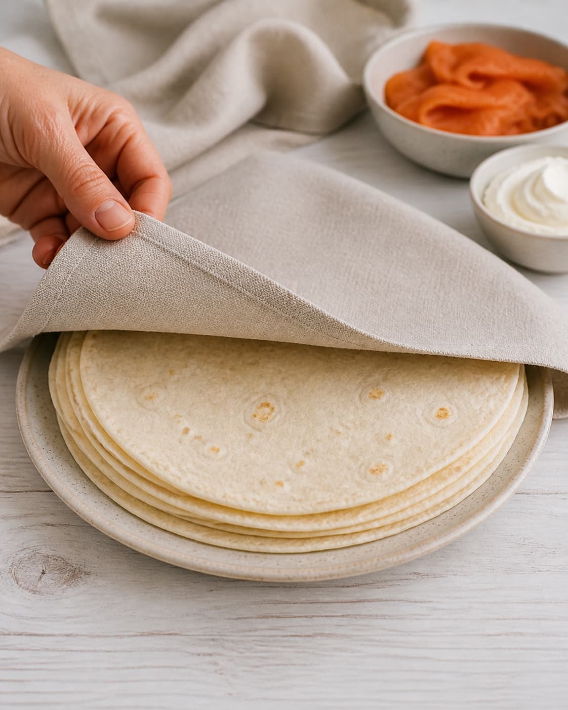 One single photorealistic instructional frame of soft flexible flatbreads warming under one cloth for Wraps med varmrøget laks, featuring tortillawraps, varmrøget laks, flødeost, with only the named equipment and no unrelated food, text, logos or packaging under tilberedningen af wraps med varmrøget laks