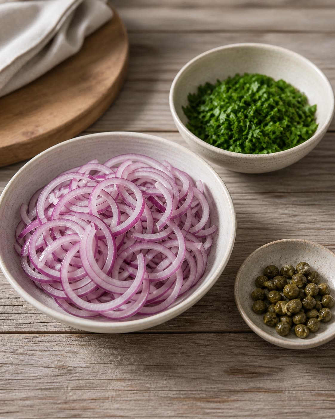 One single photorealistic instructional frame of washed and evenly cut raw ingredients arranged in separate bowls for Løgsalat, featuring rødløg, bredbladet persille, kapers, with only the named equipment and no unrelated food, text, logos or packaging under tilberedningen af løgsalat
