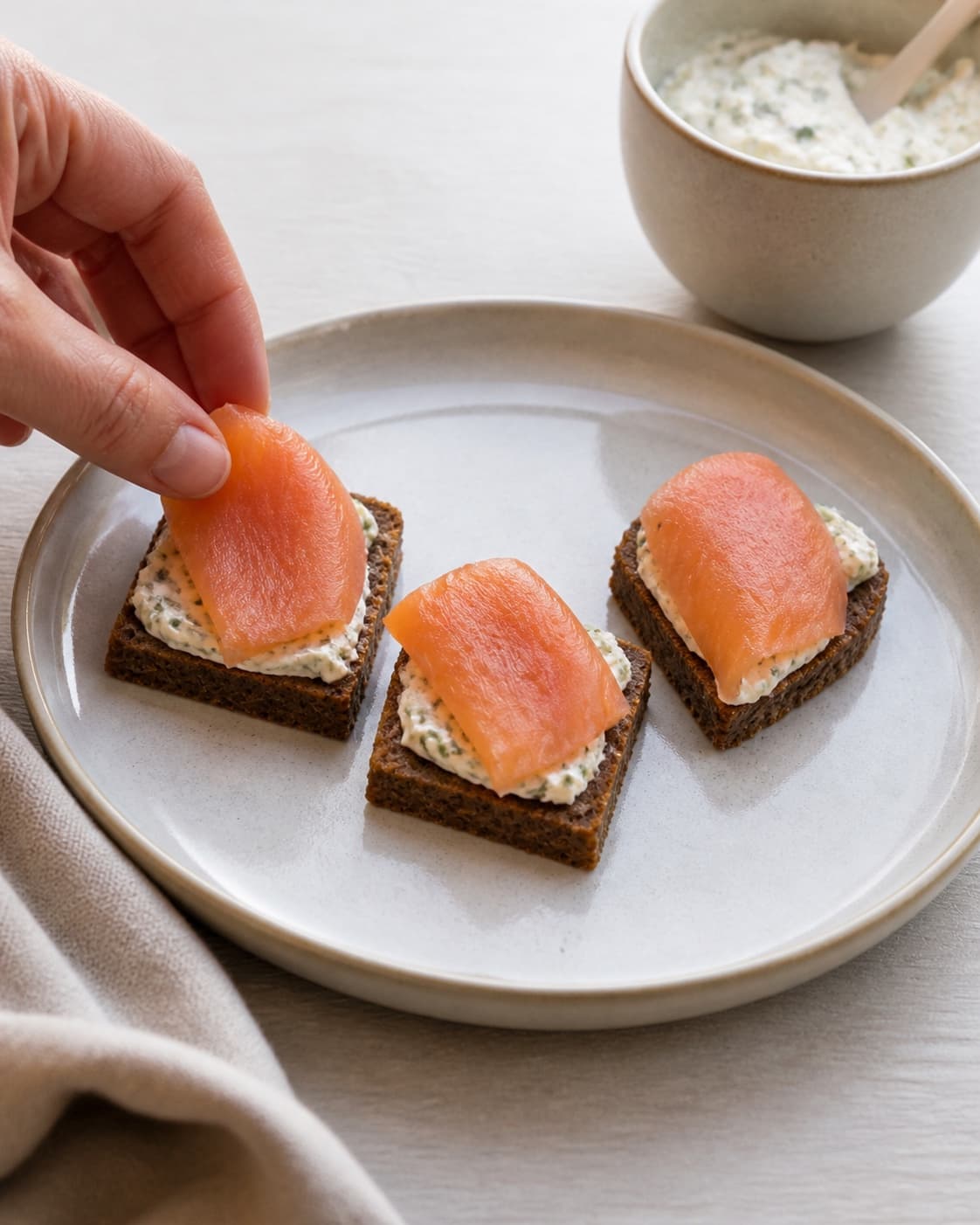 One single photorealistic instructional frame of three neat bite-size bases arranged separately for 3 nemme hapsere, featuring rugbrød, varmrøget laks, flødeost, with only the named equipment and no unrelated food, text, logos or packaging under tilberedningen af 3 nemme hapsere