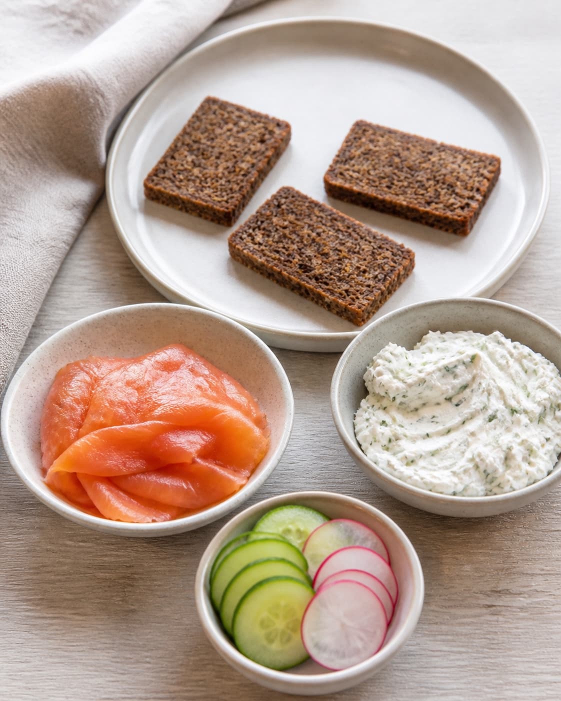 One single photorealistic instructional frame of three distinct toppings portioned in small bowls for 3 nemme hapsere, featuring rugbrød, varmrøget laks, flødeost, with only the named equipment and no unrelated food, text, logos or packaging under tilberedningen af 3 nemme hapsere