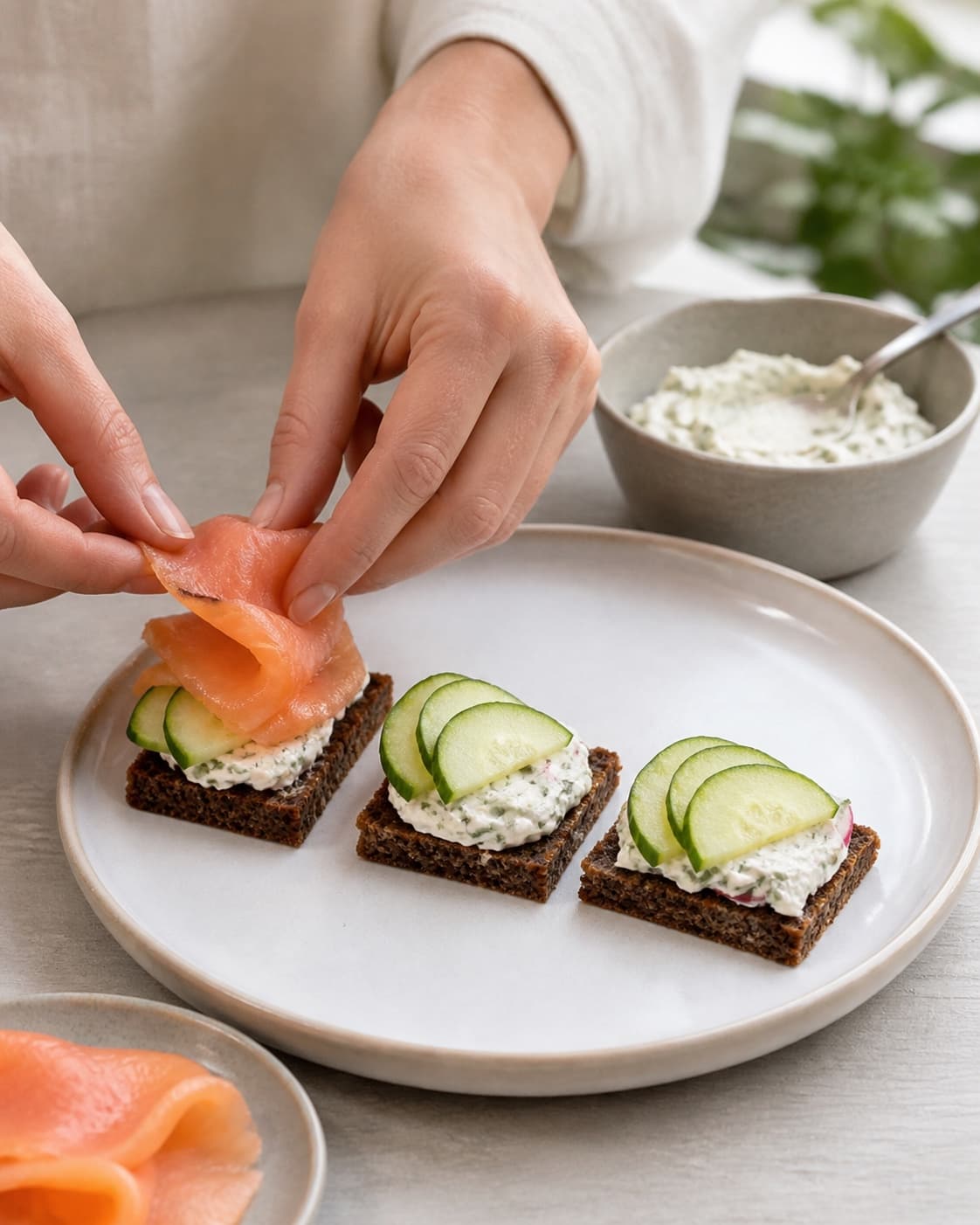 One single photorealistic instructional frame of realistic hands assembling three kinds of small bites for 3 nemme hapsere, featuring rugbrød, varmrøget laks, flødeost, with only the named equipment and no unrelated food, text, logos or packaging under tilberedningen af 3 nemme hapsere