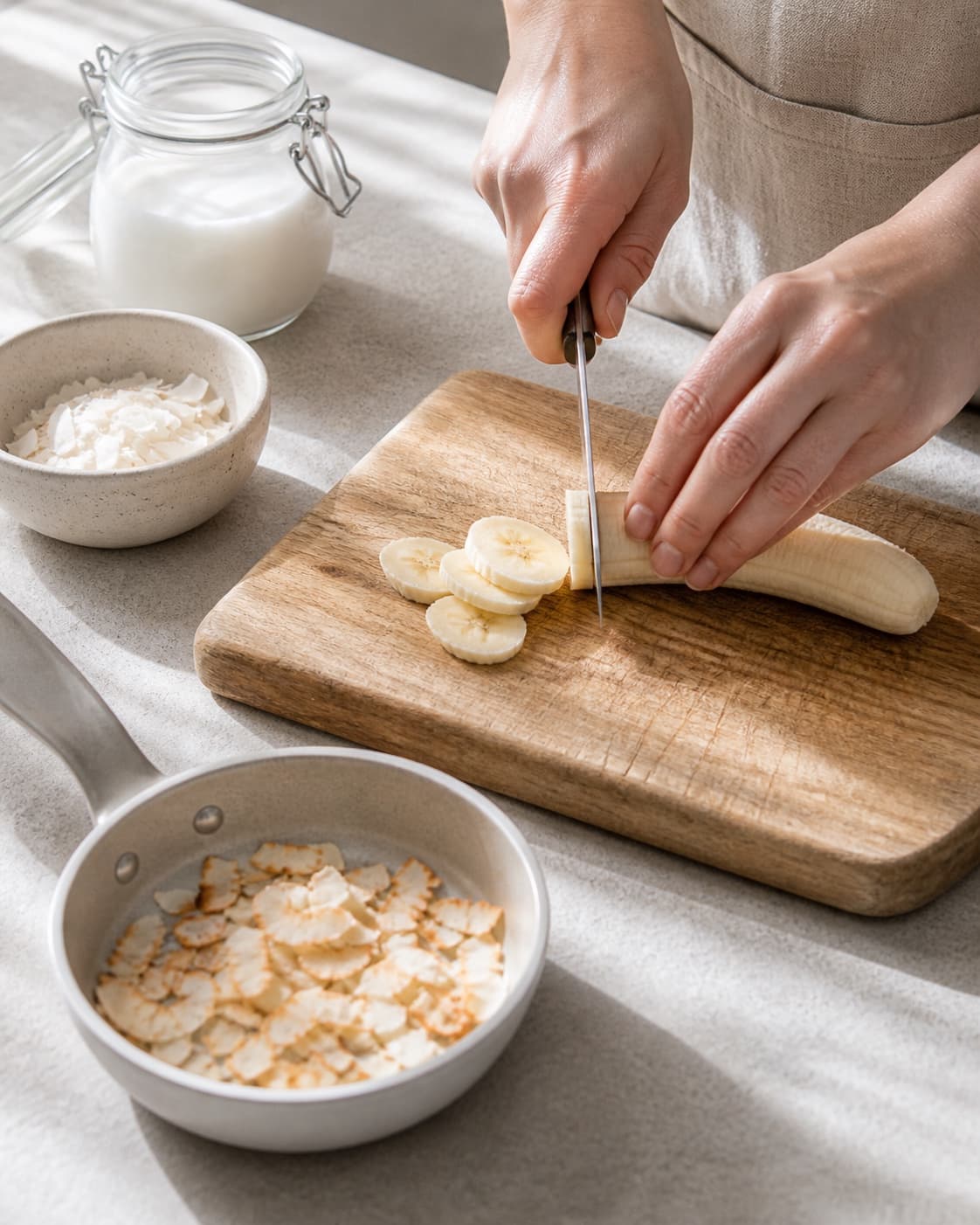 One single photorealistic instructional frame of fresh fruit being cut beside one small pan of toasted topping for Quinoagrød med kokos og banan, featuring quinoa, kokosmælk, bananer, with only the named equipment and no unrelated food, text, logos or packaging under tilberedningen af quinoagrød med kokos og banan