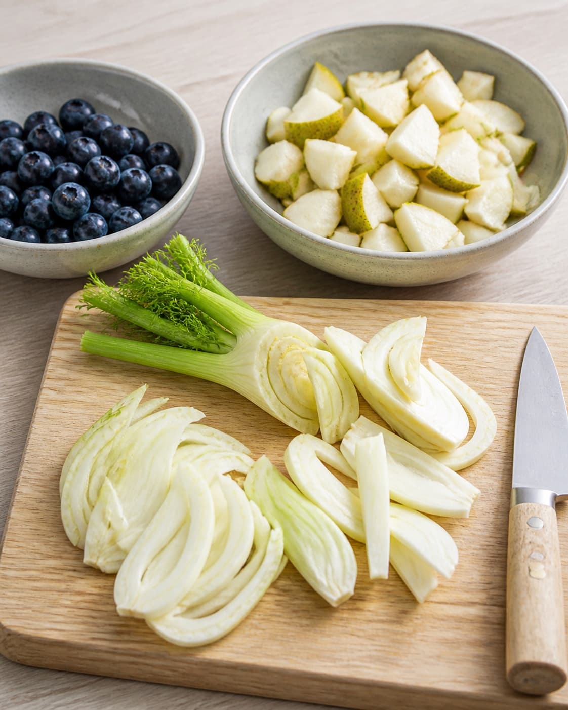 One single photorealistic instructional frame of washed and evenly cut raw ingredients arranged in separate bowls for Fennikelsalat med blåbær og pære, featuring fennikel, blåbær, pærer, with only the named equipment and no unrelated food, text, logos or packaging under tilberedningen af fennikelsalat med blåbær og pære