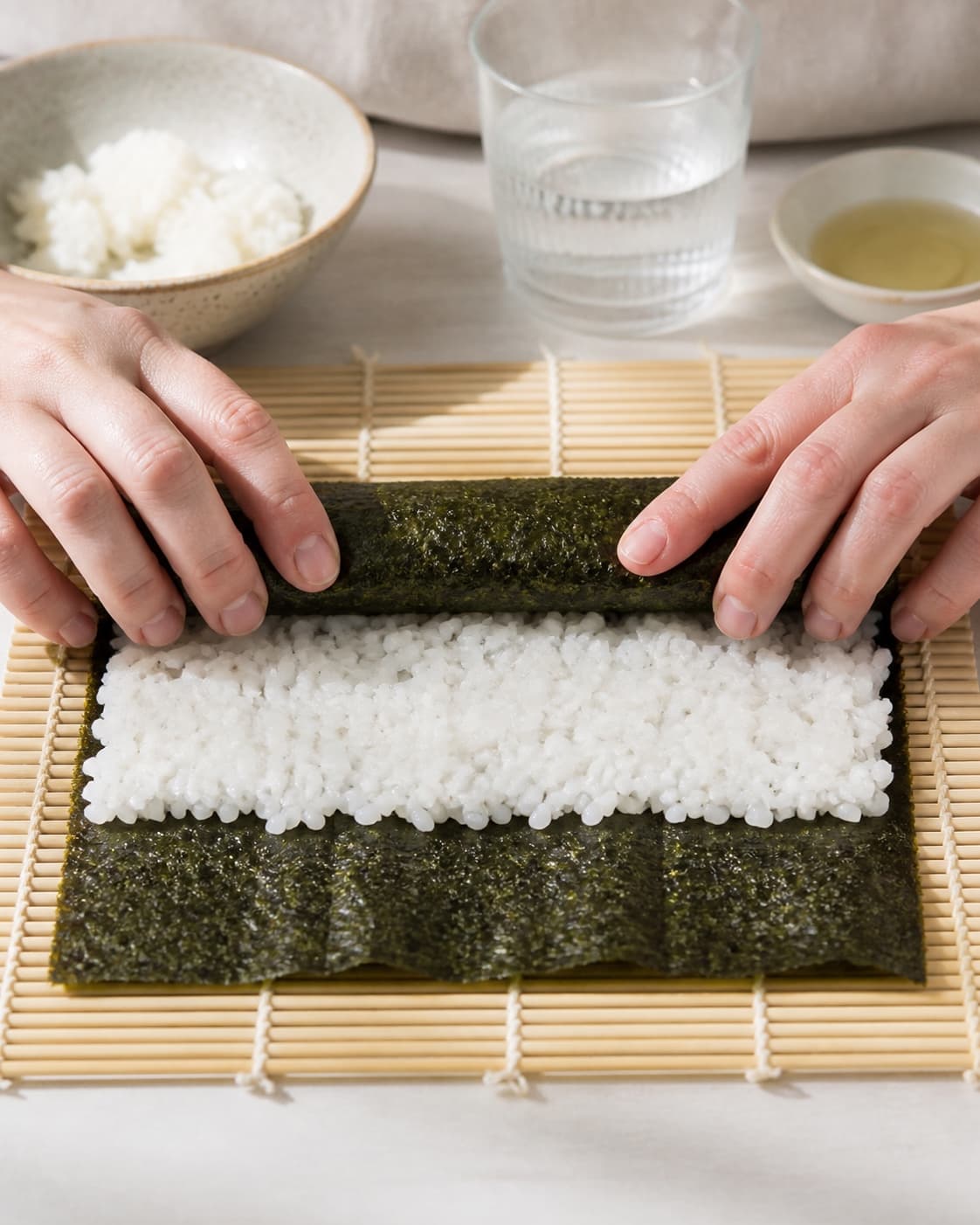 One single photorealistic instructional frame of realistic hands rolling one maki roll on a bamboo mat for Sushi i madpakken - lækre maki med laks, featuring sushiris, riseddike, noriark, with only the named equipment and no unrelated food, text, logos or packaging under tilberedningen af sushi i madpakken - lækre maki med laks