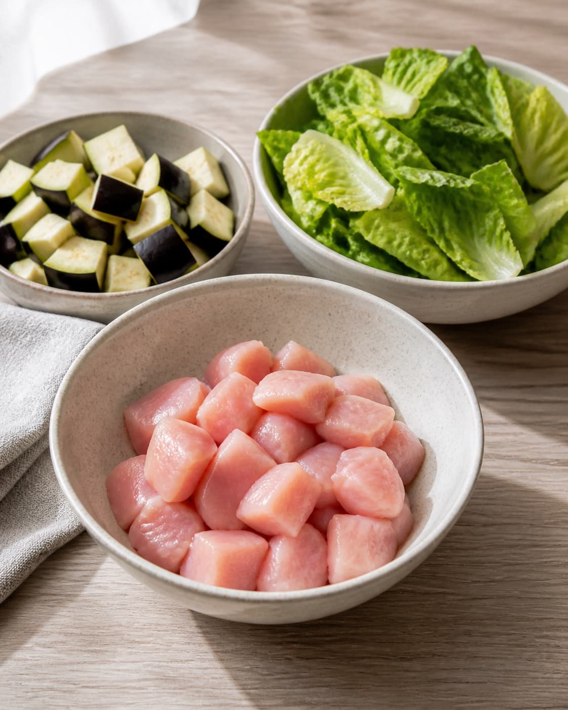 One single photorealistic instructional frame of washed and evenly cut raw ingredients arranged in separate bowls for Salat med Kylling og aubergine, featuring kyllingebryst, aubergine, romainesalat, with only the named equipment and no unrelated food, text, logos or packaging under tilberedningen af salat med kylling og aubergine