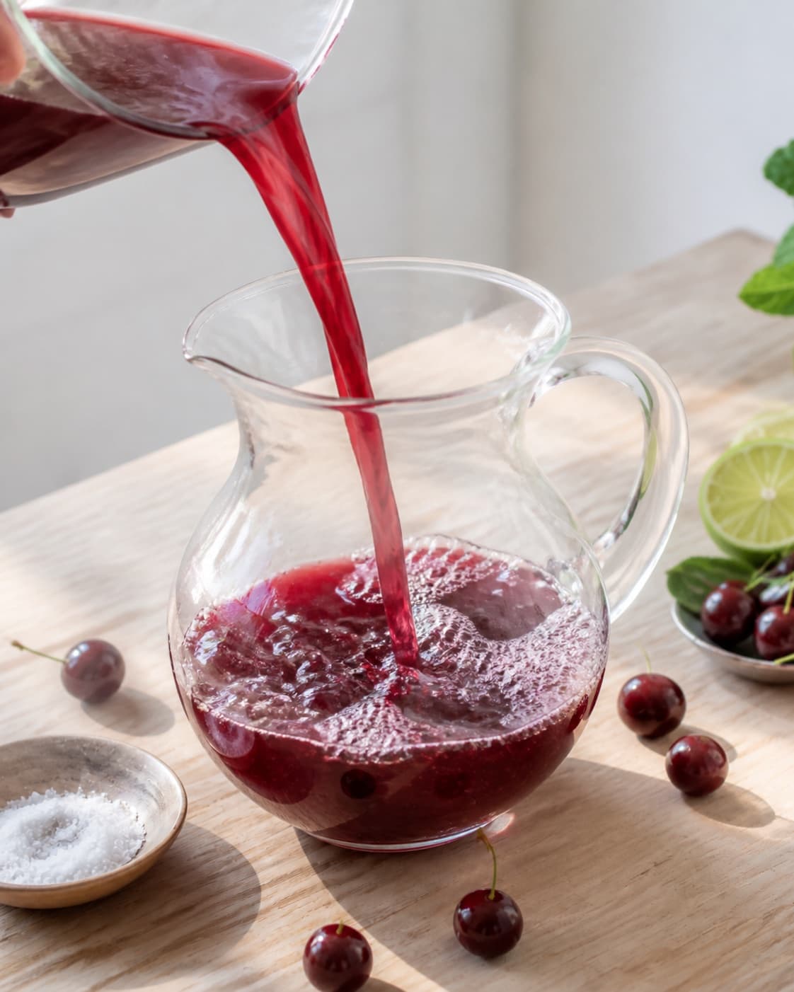 One single photorealistic instructional frame of colorful fresh juice flowing into one clean jug for Kirsebærsaft, featuring kirsebær, lime, sukker, with only the named equipment and no unrelated food, text, logos or packaging under tilberedningen af kirsebærsaft