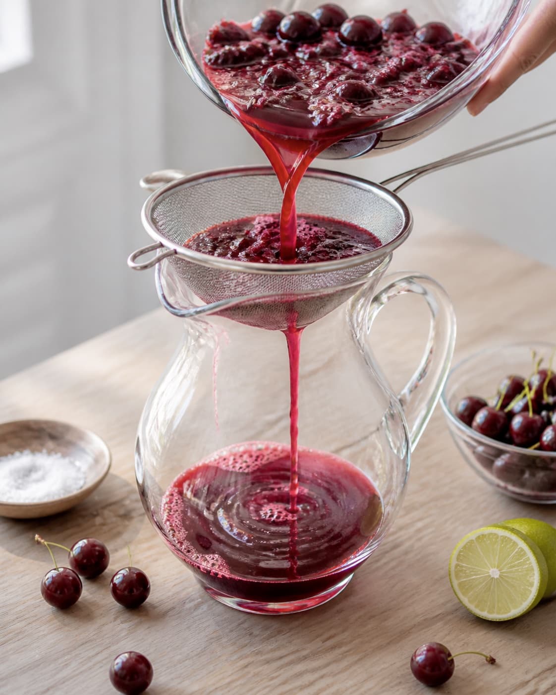 One single photorealistic instructional frame of fresh juice being strained into one jug for Kirsebærsaft, featuring kirsebær, lime, sukker, with only the named equipment and no unrelated food, text, logos or packaging under tilberedningen af kirsebærsaft