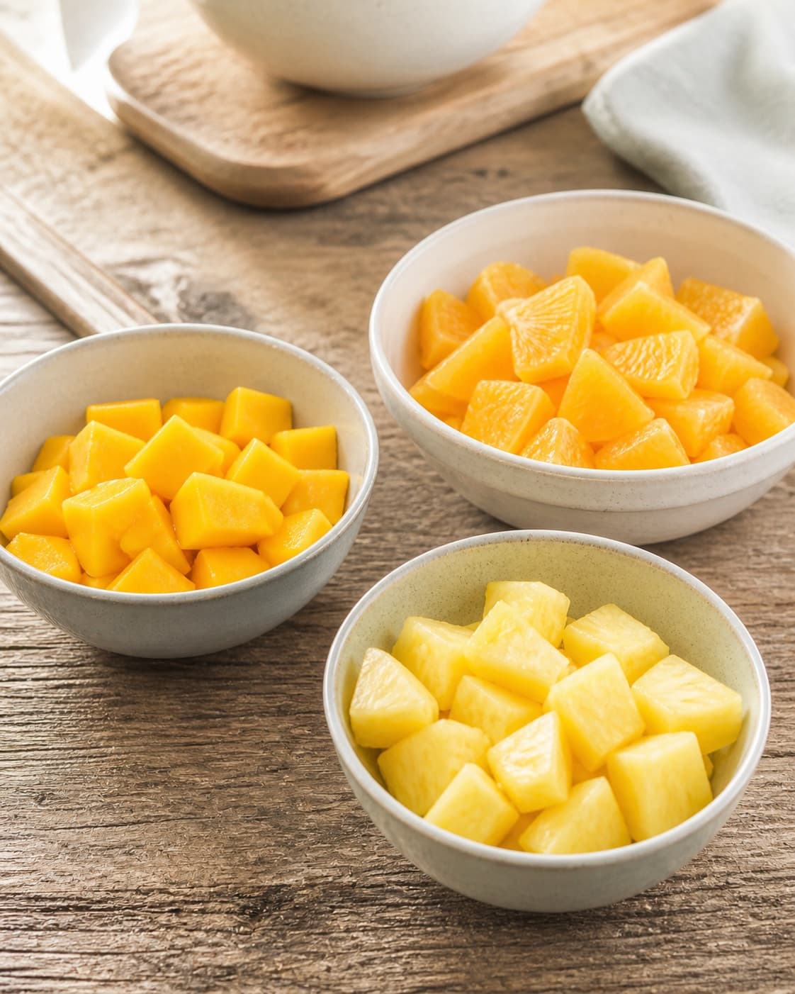 One single photorealistic instructional frame of washed and evenly cut raw ingredients arranged in separate bowls for Solskins frugtsalat, featuring ananas, mango, appelsin, with only the named equipment and no unrelated food, text, logos or packaging under tilberedningen af solskins frugtsalat