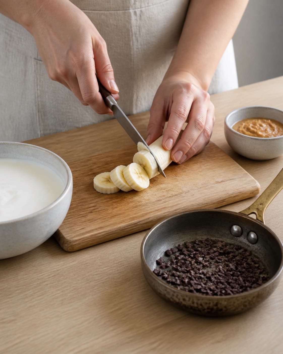 One single photorealistic instructional frame of fresh fruit being cut beside one small pan of toasted topping for Frugtyoghurt med banan, peanutbutter og kakaonibs, featuring yoghurt naturel, banan, peanutbutter, with only the named equipment and no unrelated food, text, logos or packaging under tilberedningen af frugtyoghurt med banan, peanutbutter og kakaonibs
