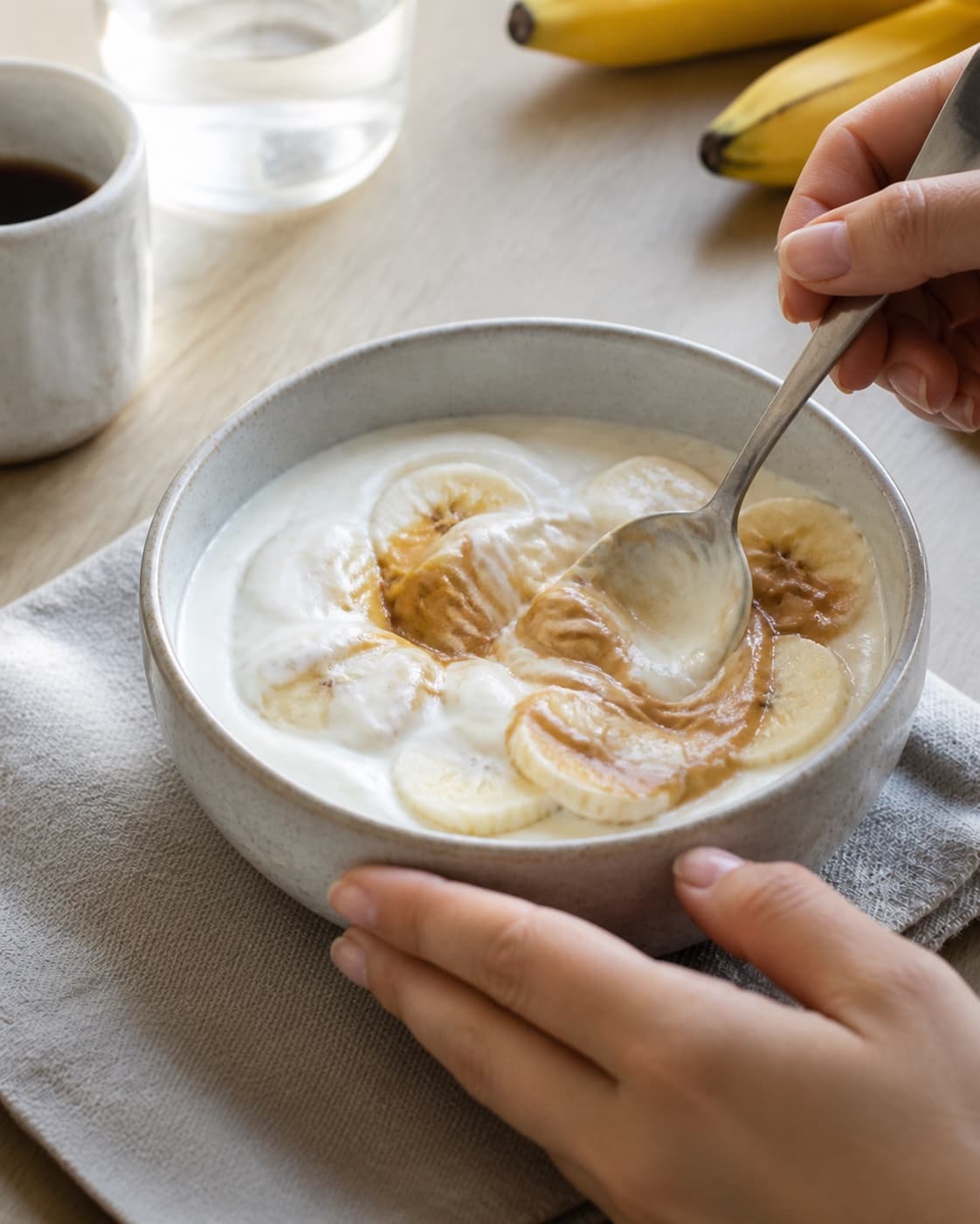 One single photorealistic instructional frame of the porridge consistency being adjusted with a spoon for Frugtyoghurt med banan, peanutbutter og kakaonibs, featuring yoghurt naturel, banan, peanutbutter, with only the named equipment and no unrelated food, text, logos or packaging under tilberedningen af frugtyoghurt med banan, peanutbutter og kakaonibs