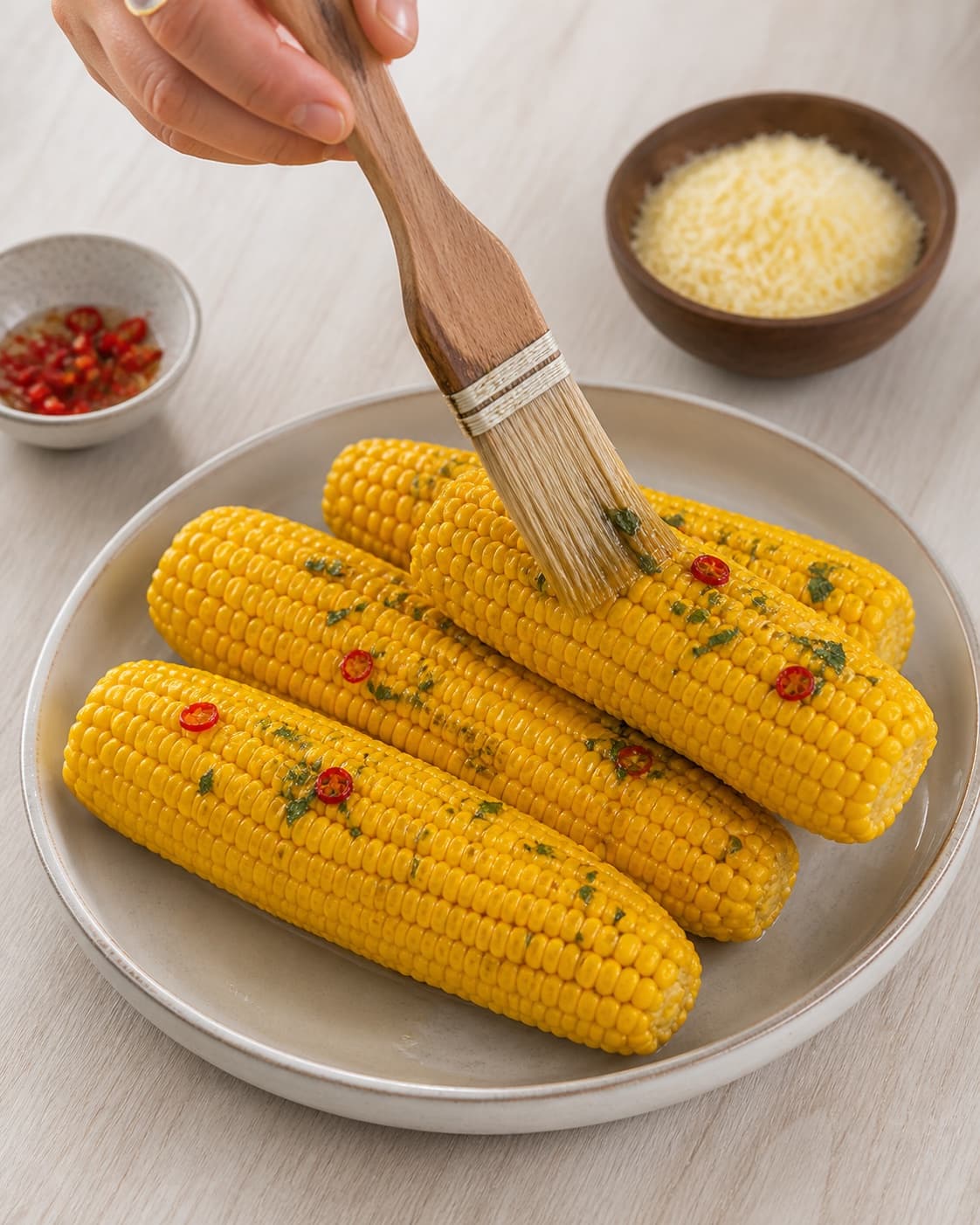 One single photorealistic instructional frame of the main ingredient being brushed with seasoning for Majskolber med parmesan og chili, featuring majskolber, parmesan, chili, with only the named equipment and no unrelated food, text, logos or packaging under tilberedningen af majskolber med parmesan og chili