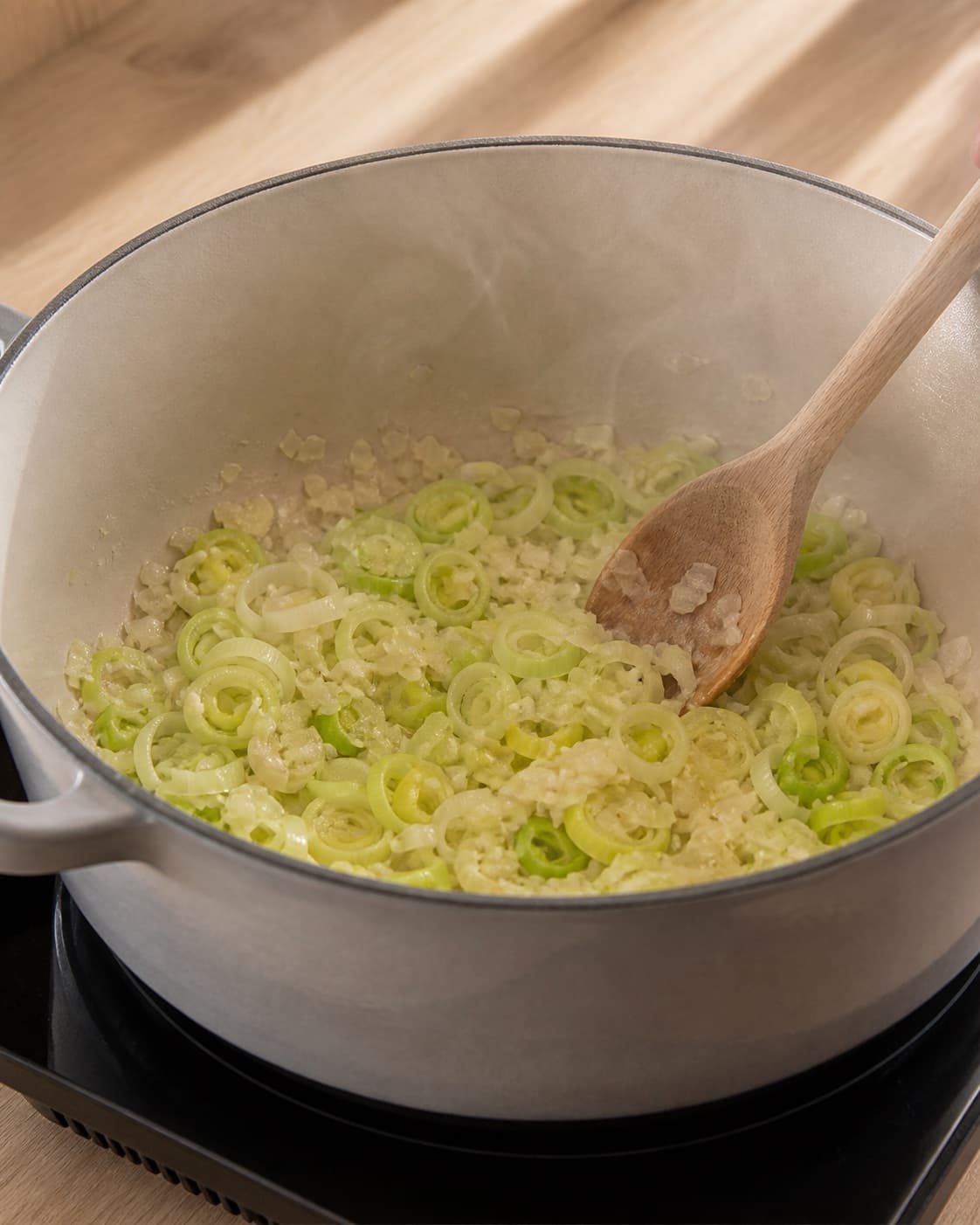One single photorealistic instructional frame of aromatic vegetables softening in the same heavy pot for Bygotto med varmrøget laks, featuring perlebyg, varmrøget laks, porrer, with only the named equipment and no unrelated food, text, logos or packaging under tilberedningen af bygotto med varmrøget laks