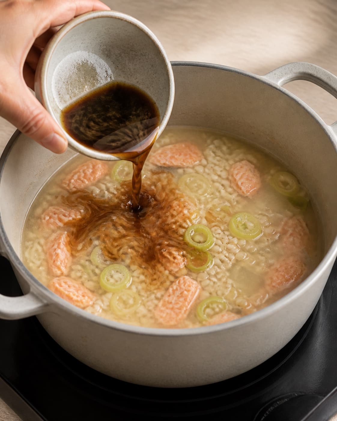 One single photorealistic instructional frame of dark fond dissolving into the added cooking liquid for Bygotto med varmrøget laks, featuring perlebyg, varmrøget laks, porrer, with only the named equipment and no unrelated food, text, logos or packaging under tilberedningen af bygotto med varmrøget laks