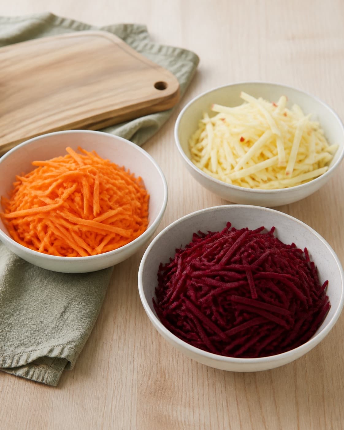 One single photorealistic instructional frame of washed and evenly cut raw ingredients arranged in separate bowls for Råkost med rødbede, featuring rødbede, gulerødder, æble, with only the named equipment and no unrelated food, text, logos or packaging under tilberedningen af råkost med rødbede
