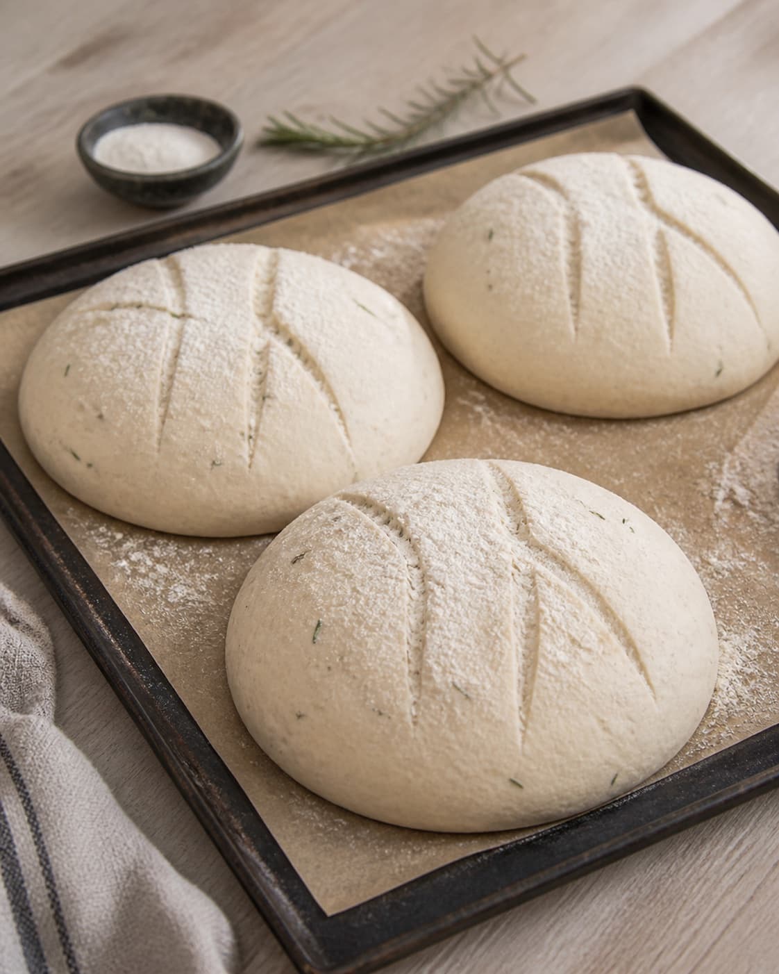 One single photorealistic instructional frame of shaped pale dough pieces visibly risen on one baking tray for Brød med rosmarin, featuring hvedemel, gær, frisk rosmarin, with only the named equipment and no unrelated food, text, logos or packaging under tilberedningen af brød med rosmarin