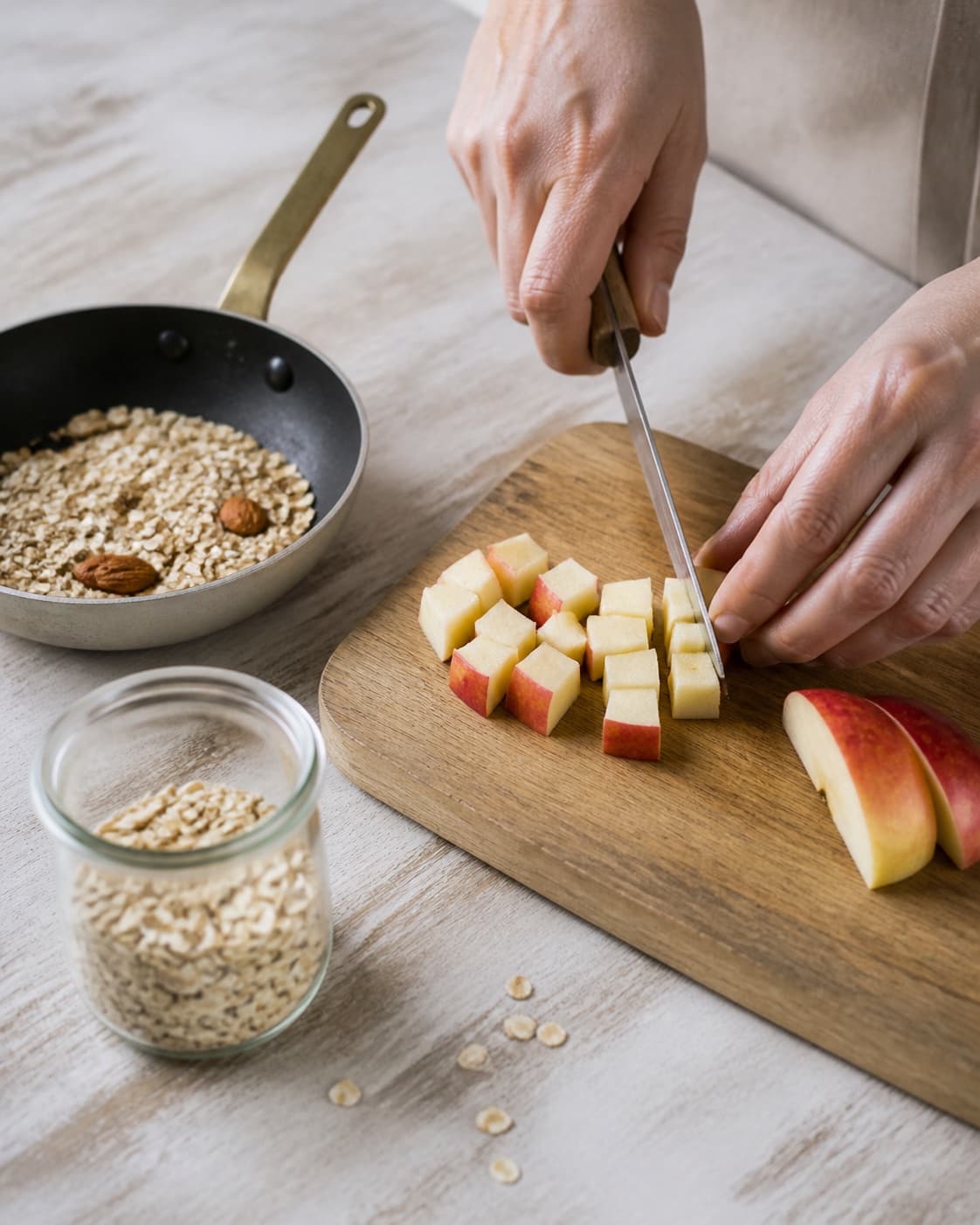 One single photorealistic instructional frame of fresh fruit being cut beside one small pan of toasted topping for Drænet yoghurt med knas og æbler, featuring græsk yoghurt, æbler, havregryn, with only the named equipment and no unrelated food, text, logos or packaging under tilberedningen af drænet yoghurt med knas og æbler