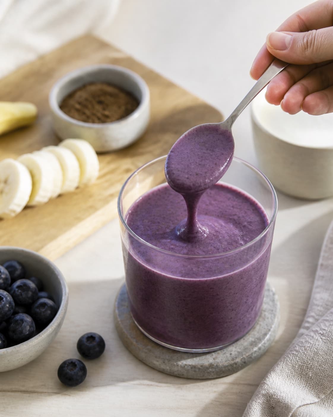 One single photorealistic instructional frame of one spoon checking the thick smoothie consistency for Blåbær og lakridssmoothie, featuring blåbær, banan, lakridspulver, with only the named equipment and no unrelated food, text, logos or packaging under tilberedningen af blåbær og lakridssmoothie