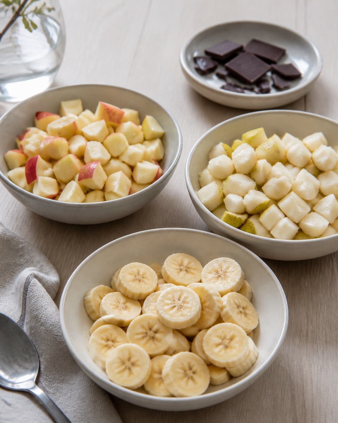 One single photorealistic instructional frame of washed and evenly cut raw ingredients arranged in separate bowls for Frugtsalat med chokolade, featuring æbler, pærer, banan, with only the named equipment and no unrelated food, text, logos or packaging under tilberedningen af frugtsalat med chokolade