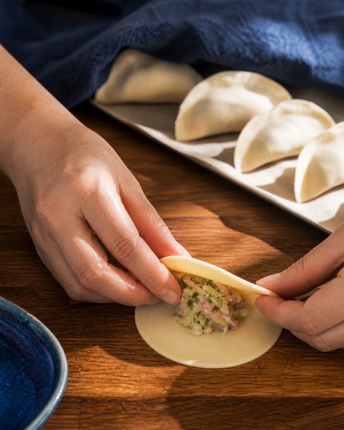 To hænder folder en rund dumplingdej over en lille portion koldt svinekøds- og kålfyld ved forseglede rå halvmåner på papir