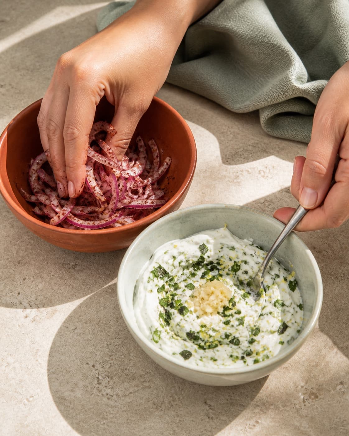 To hænder masserer rødløg med sumak i terracottaskål og rører en separat tyk yoghurt med mynte, citron og hvidløg