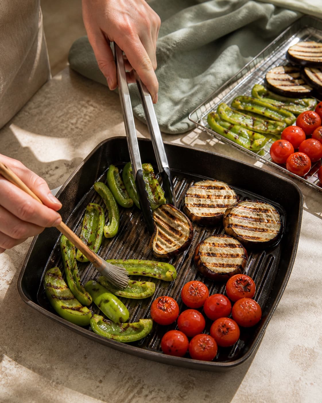 To hænder pensler og vender aubergine, grøn peber og cherrytomater i adskilte zoner på grillpanden ved mørke striber