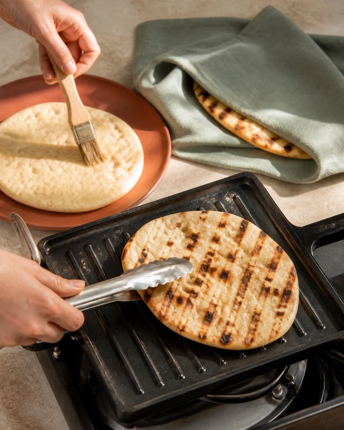 To hænder pensler ét fladbrød ganske let med vand og vender et andet kort på grillen ved et løst dækket varmt brød