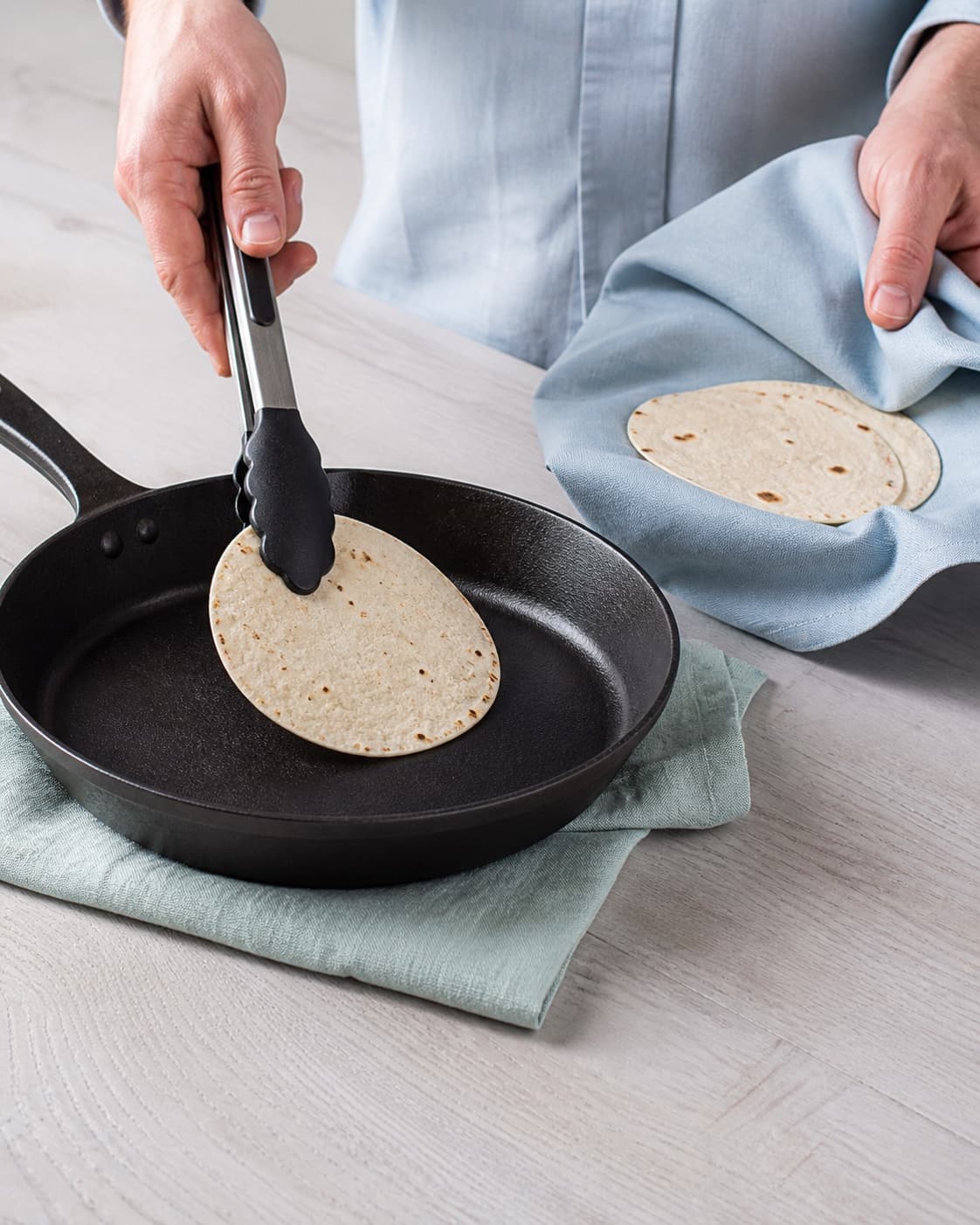 To hænder vender en lille tortilla på tør varm pande og holder tidligere varme smidige tortillas løst i et rent lyseblåt klæde