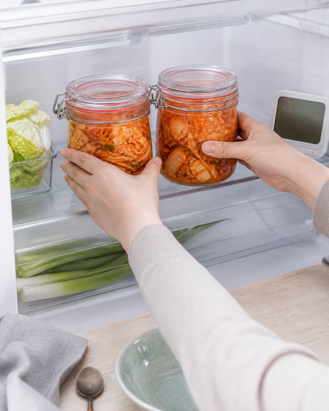 To hænder sætter lukkede glas med frisk hurtig kimchi direkte på en kold køleskabshylde ved et ulæseligt termometer